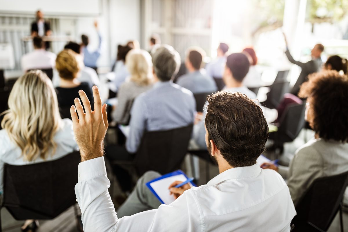 Rear view of a businessman raising his hand to ask the question on a seminar with large group of his colleagues.