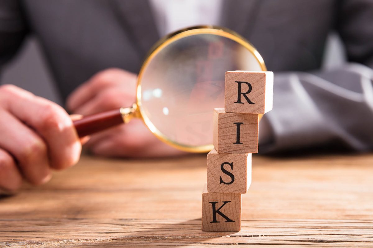 Close-up Of Wooden Blocks With Risk Word In Front Of Businessperson's Hand Holding Magnifying Glass